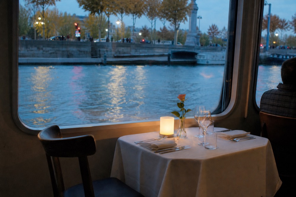 A candlelit dinner table set beside the window of a Seine cruise boat, no text, no logos, no people