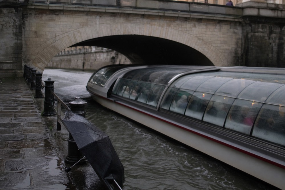 A covered Seine cruise boat cruising under a stone bridge in soft rain, no text, no logos, no people.