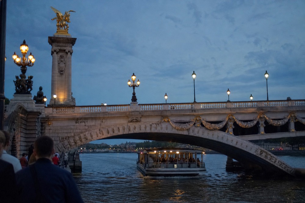 A cruise boat passing under one of Paris's ornate illuminated bridges at dusk, no text, no logos, no people