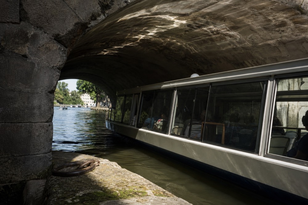 A large glass-sided sightseeing boat passing under a stone bridge on the Seine, no text, no logos, no people