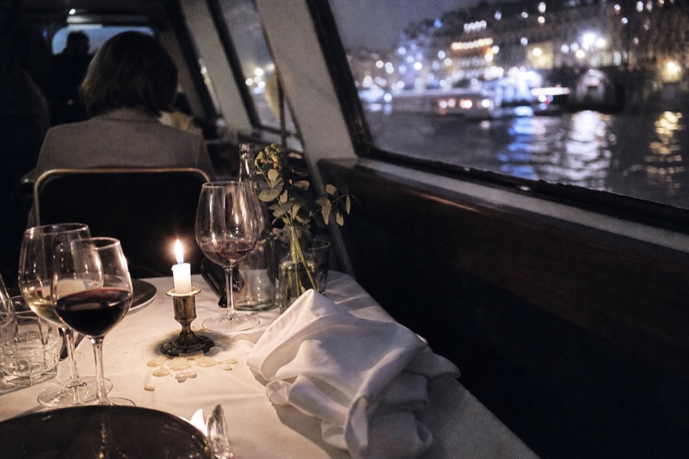 A candlelit dinner table set with wine glasses on a boat with Paris lights outside the window, no text, no logos, no people