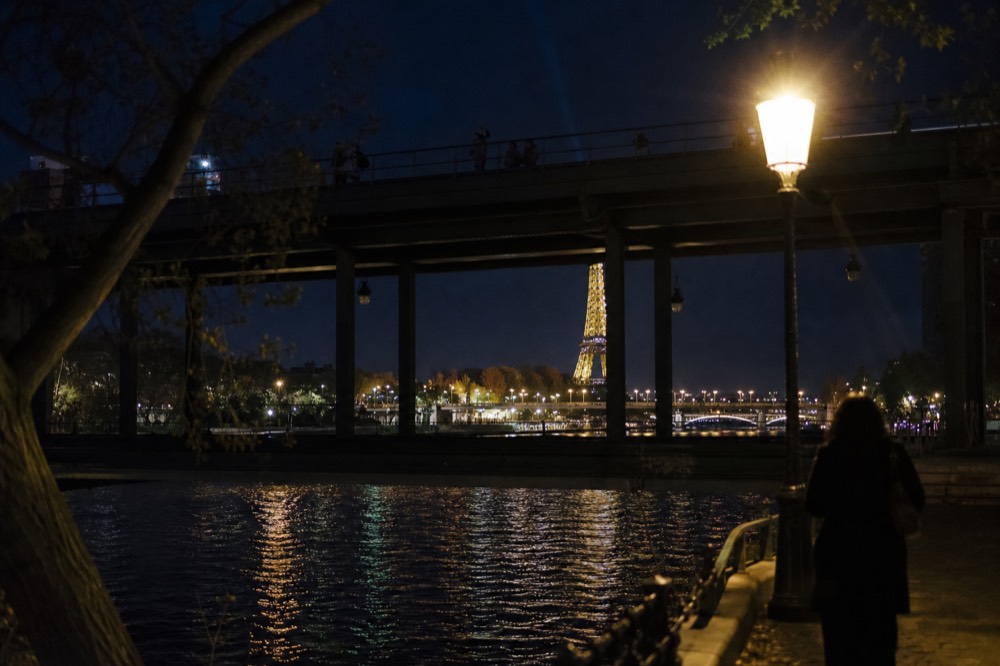 The Eiffel Tower mid-sparkle seen from the Seine with a bridge silhouette in the foreground, no text, no logos, no people