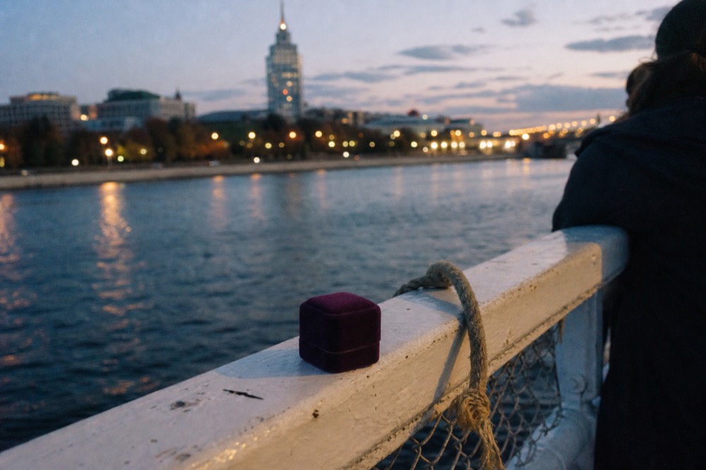 A ring box resting on a boat railing with the Seine and city lights blurred behind, no text, no logos, no people