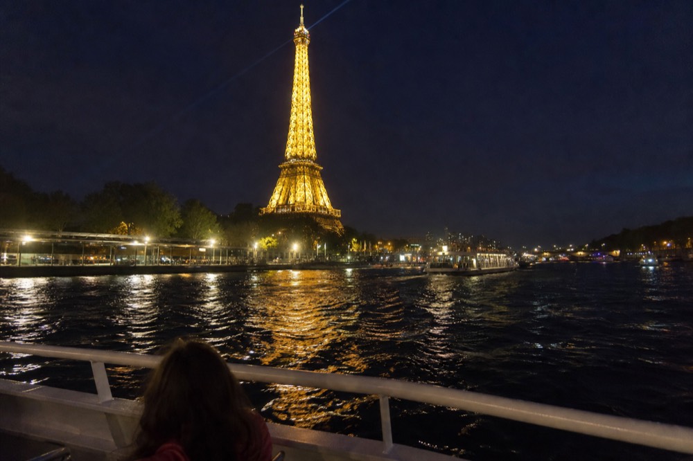 The illuminated Eiffel Tower seen from the water with a cruise boat railing in the foreground, no text, no logos, no people