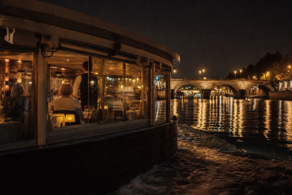 A dinner cruise boat with lit interior tables gliding past illuminated Paris bridges, no text, no logos, no people