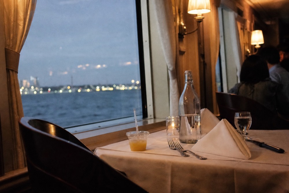 Laid dinner table beside a wide window on a river boat, city lights outside, no text, no logos, no people.