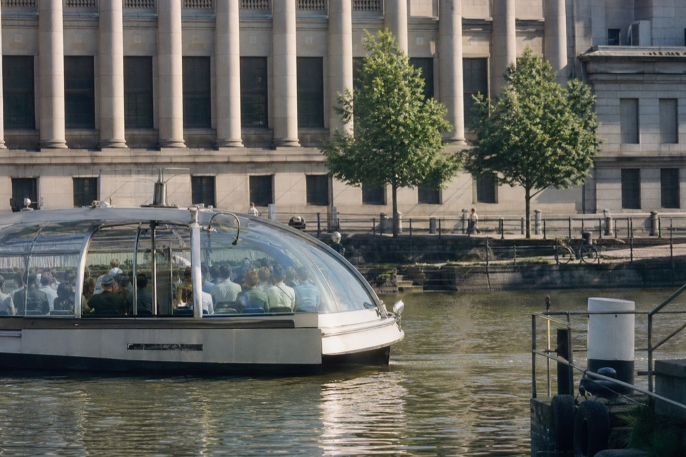 Glass-enclosed shuttle boat approaching a Seine stop with a terrace of seated passengers, no text, no logos, no people.