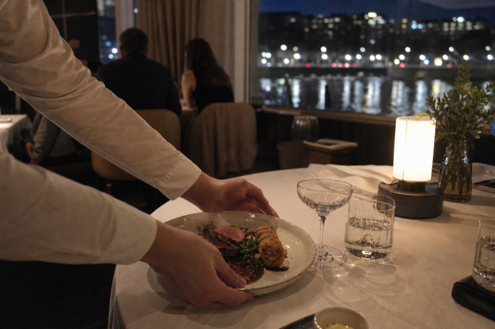 Waiter serving a plated gourmet main course inside a Seine dinner cruise dining room, no text, no logos, no people