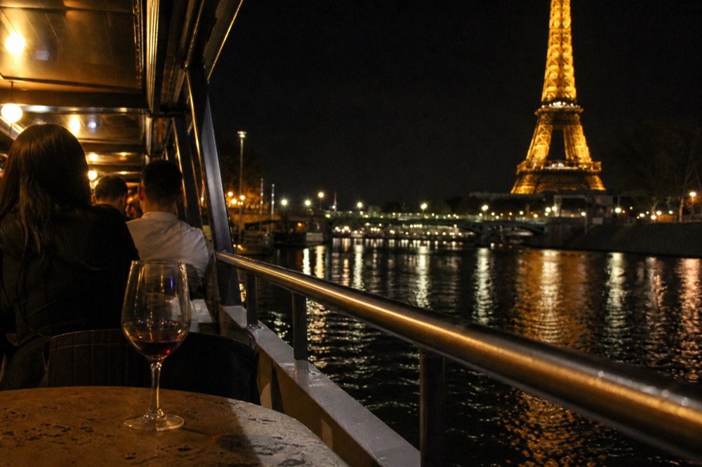 Dinner cruise boat deck lit at night with the sparkling Eiffel Tower reflected in the Seine, no text, no logos, no people