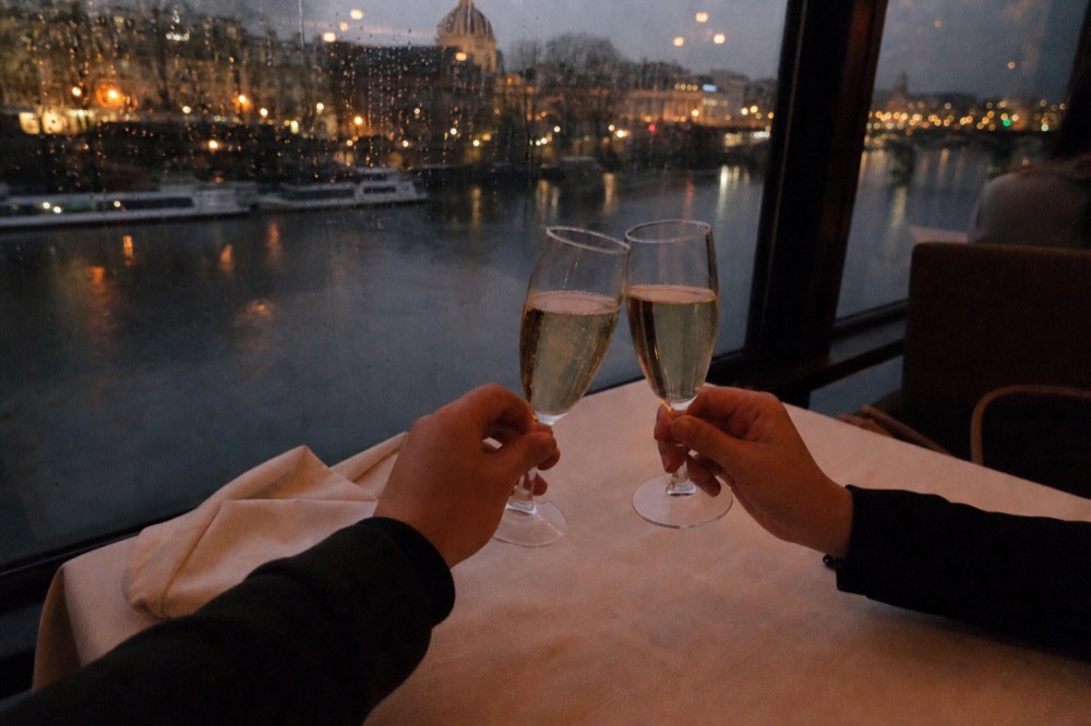 Couple toasting with Champagne glasses at a window table as Paris lights pass outside, no text, no logos, no people